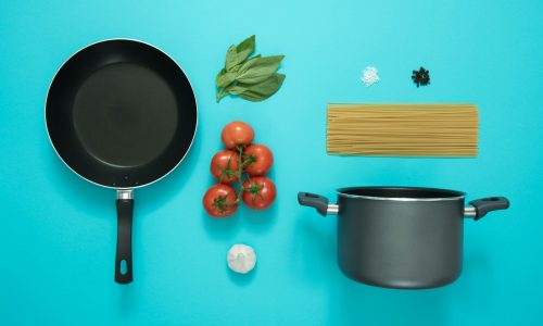 Frying pan, pot, tomatoes, spaghetti, basil, garlic, salt, and pepper on a blue background for a cooking theme